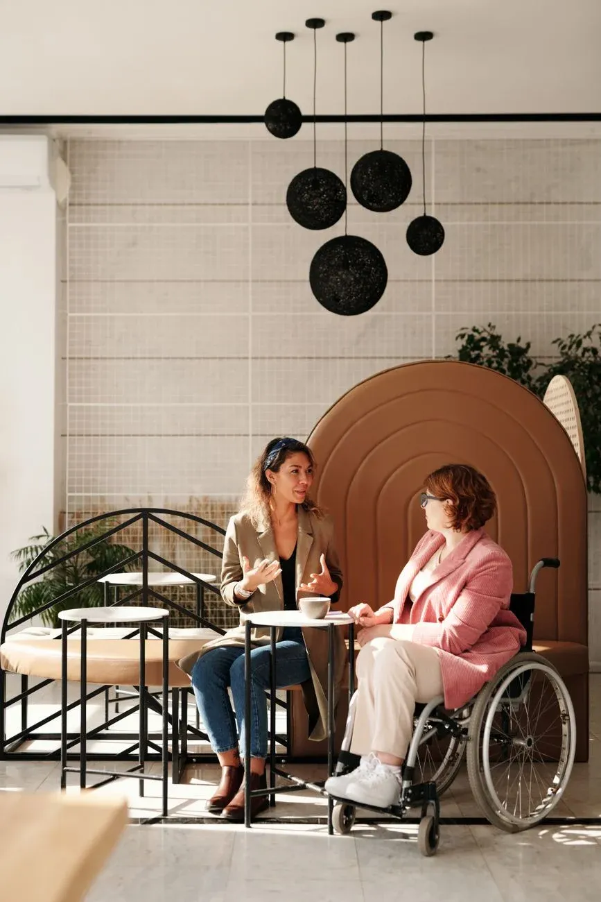 Two women in conversation, one using a wheelchair, in a sunlit cafe